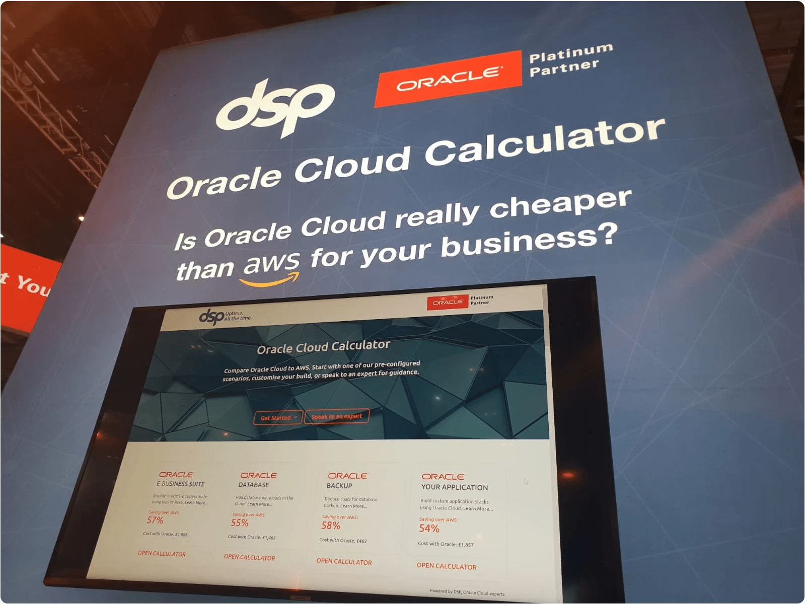 A photo of the DSP exhibition stand at Oracle OpenWorld Europe. A large screen displays the Oracle Cloud Calculator website. The screen sits inside a large exhibition stand with the words, 'Oracle Cloud Calculator. Is Oracle Cloud really cheaper than AWS for your business?
